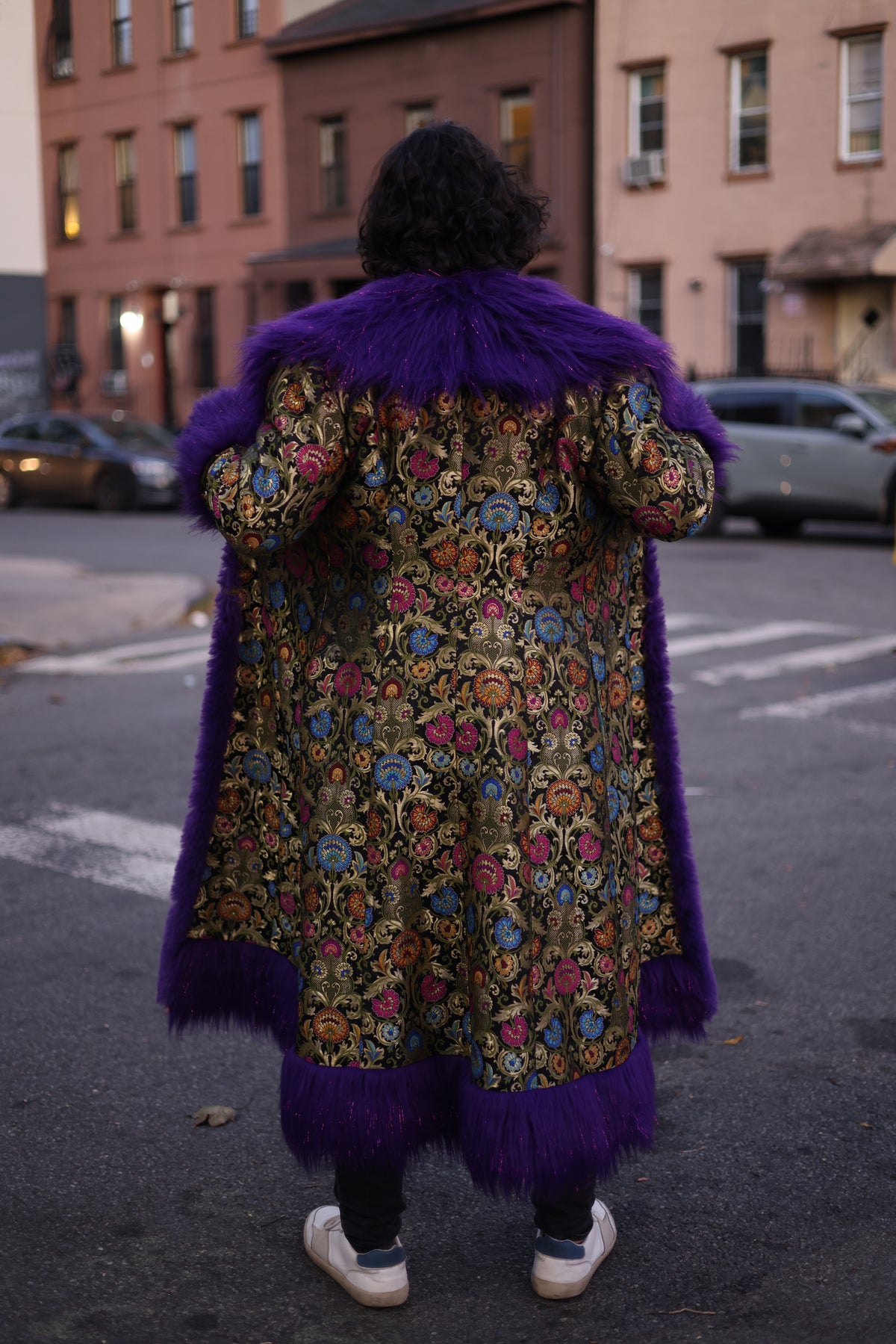 Back view of a model wearing the PURPLE full-length silk brocade coat with intricate multicolor floral patterns, an oversized purple faux fur collar, and matching hem trim.